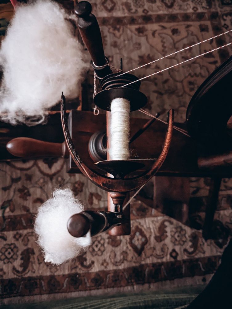 A moody and dark photo of a flyer from a 1700s flax wheel. On it are two puffs of natural white wool. The bobbin has a decent amount of wool spun on it. 