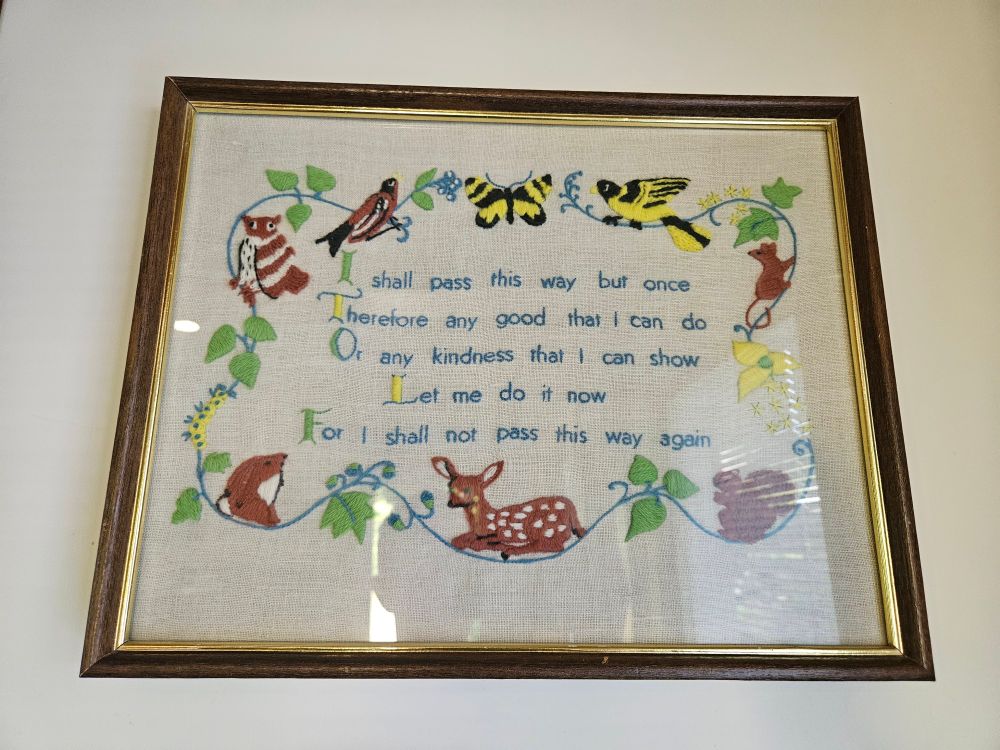 An old framed needlework made by Tristan's grandmother, with a border of foliage and charmingly wonky woodland creatures. In the centre it reads: "I shall pass this way but once / Therefore any good that I can do / Or any kindness that I can show / Let me do it now / For I shall not pass this way again."