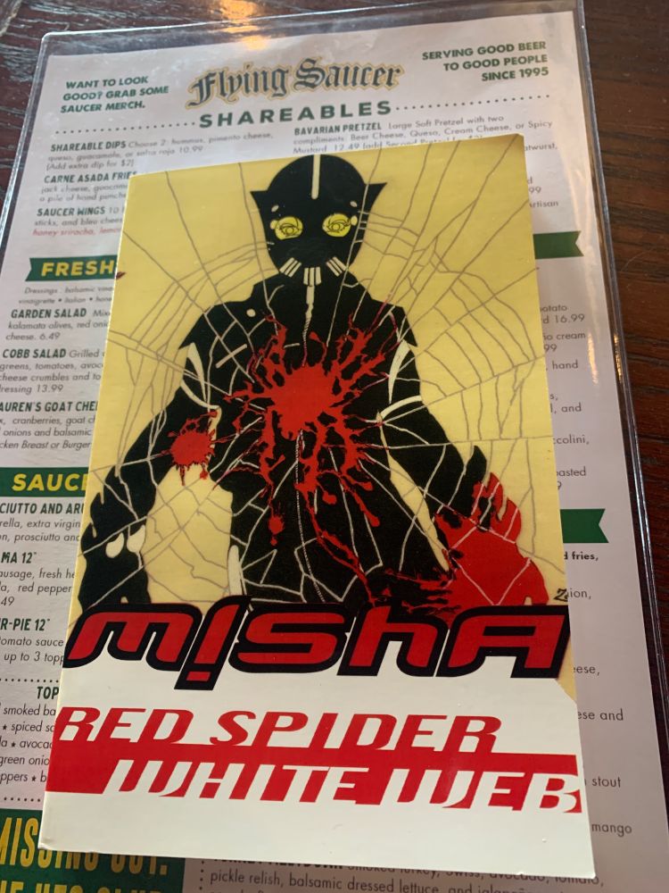 A copy of “Red Spider, White Web” by Misha Nogha, cover by Joey Zone (published by Wordcraft of Oregon), atop a menu for The Flying Saucer pub.