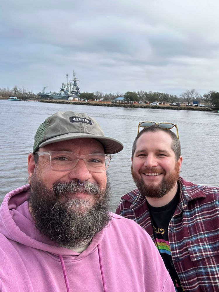 Two bearded men in love, in front of th the Cape Fear River with The Battleship NC in the background. 
