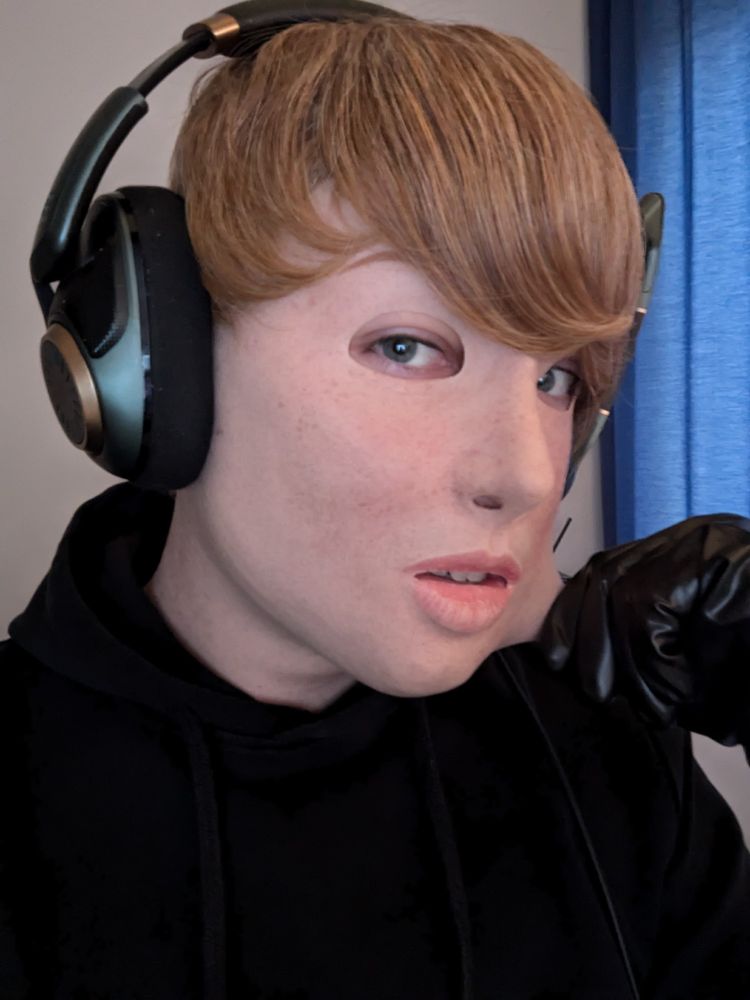 A portrait of a person wearing a realistic silicone mask of a freckled Girl with short blonde Hair, wearing a black hoodie tugging at the mask with a hand in a black latex glove.