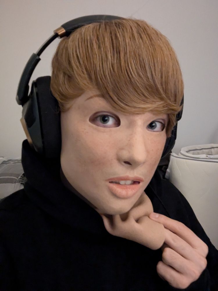 Portrait of a person wearing a realistic silicone mask of a light-skinned freckled girl with a short blonde wig wearing headphones and a black hoodie, tugging at the edge of the mask.