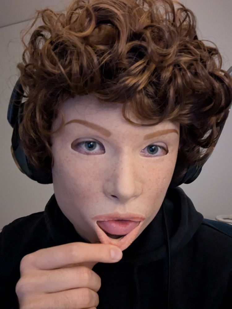 A person wearing a realistic silicone mask of a light-skinned freckled girl with a short, curly, light brown wig. They are pulling at the lower lip of the mask, revealing their own lip.