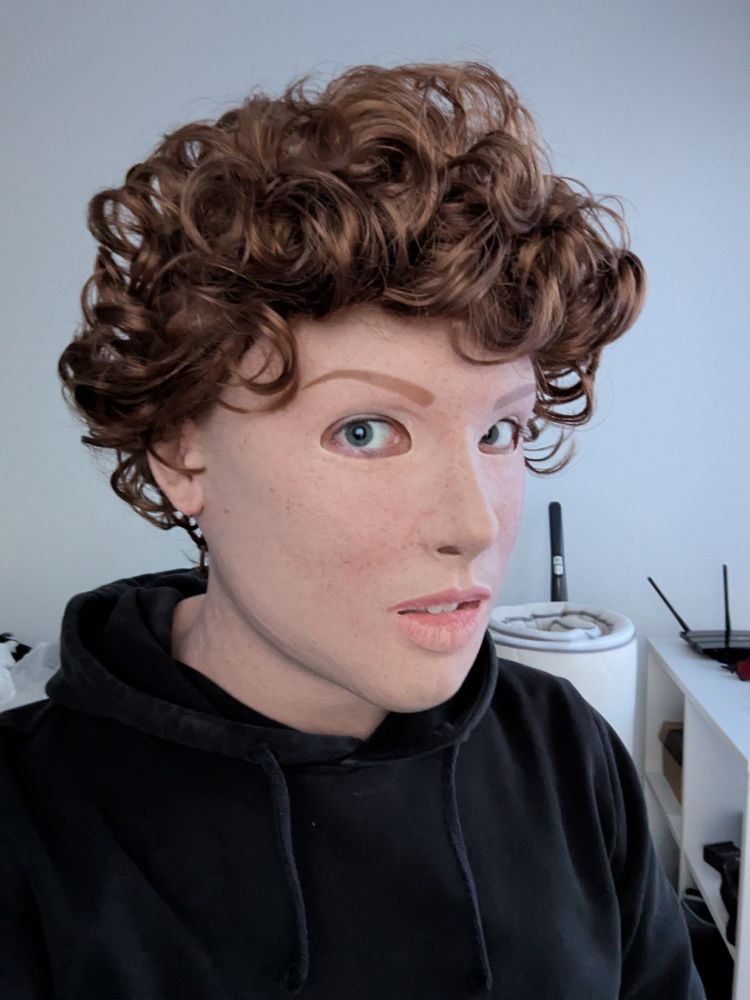 A portrait of a person wearing a realistic silicone mask of a light-skinned freckled girl with a short, curly, light brown wig.