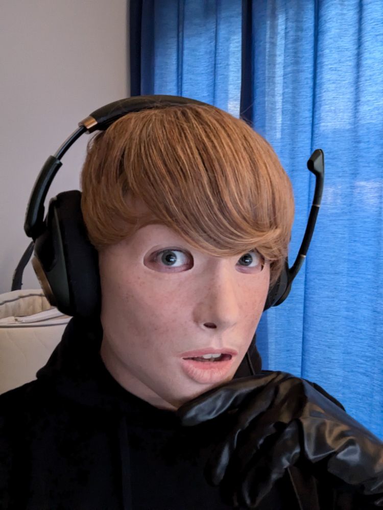 A portrait of a person wearing a realistic silicone mask of a freckled Girl with short blonde Hair, wearing a black hoodie and holding a hand in a black latex glove to the face.