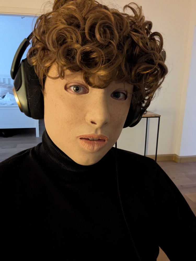 A portrait of a person wearing a realistic silicone mask of a light-skinned freckled girl. The person is also wearing a curly dark-blonde wig and a gaming headset.