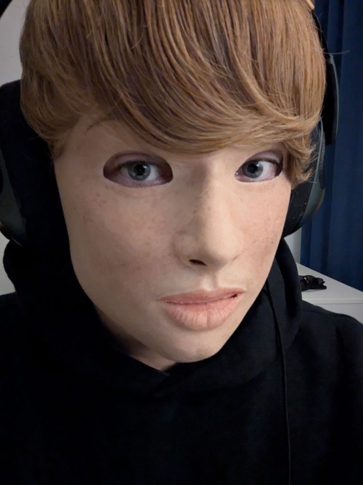Portrait of a person wearing a realistic silicone mask of a light-skinned freckled girl with a short blonde wig wearing headphones and a black hoodie.