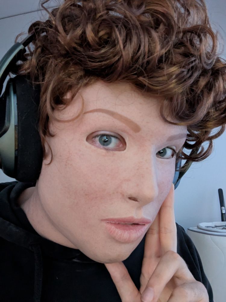 A close-up of a person wearing a realistic silicone mask of a light-skinned freckled girl with a short, curly, light brown wig.