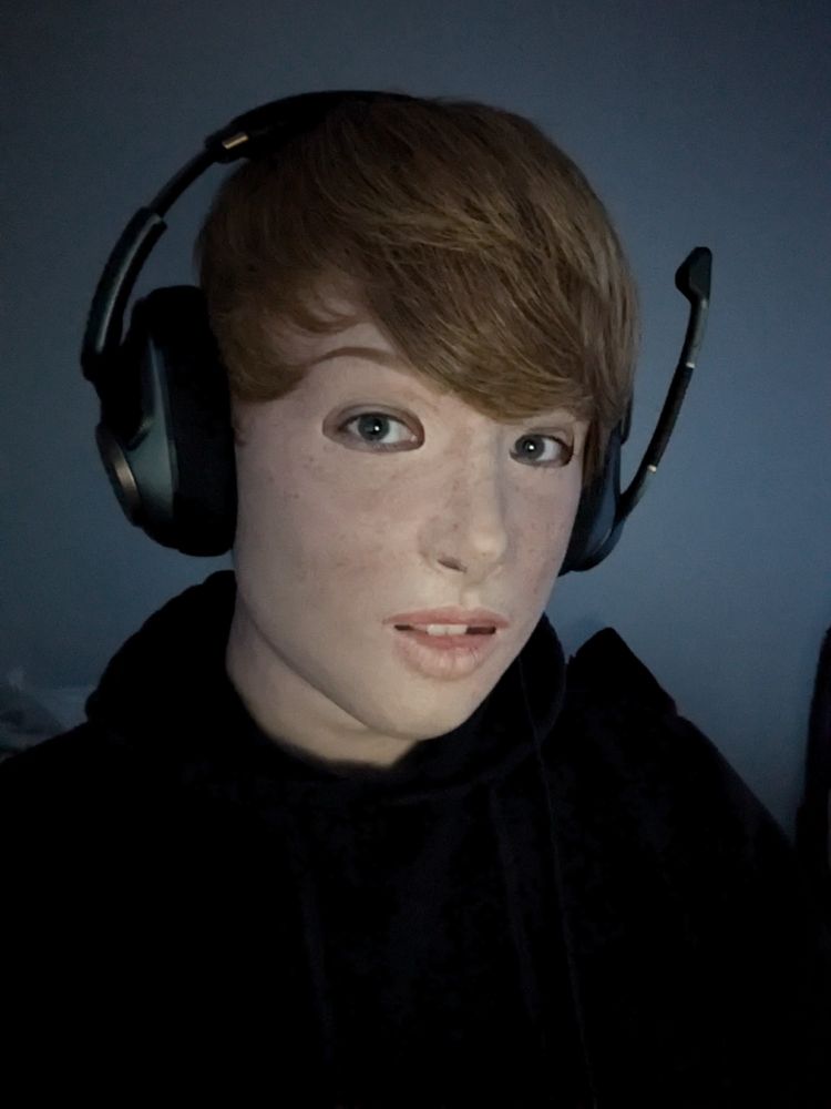A portrait of a person in a realistic silicone mask of a light-skinned Girl with short blonde hair, wearing a black hoodie and a Gaming-Headset in a dark room.