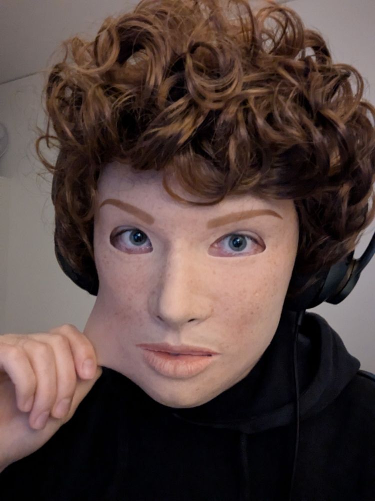 A person wearing a realistic silicone mask of a light-skinned freckled girl with a short, curly, light brown wig. They are pulling the cheek of the mask.