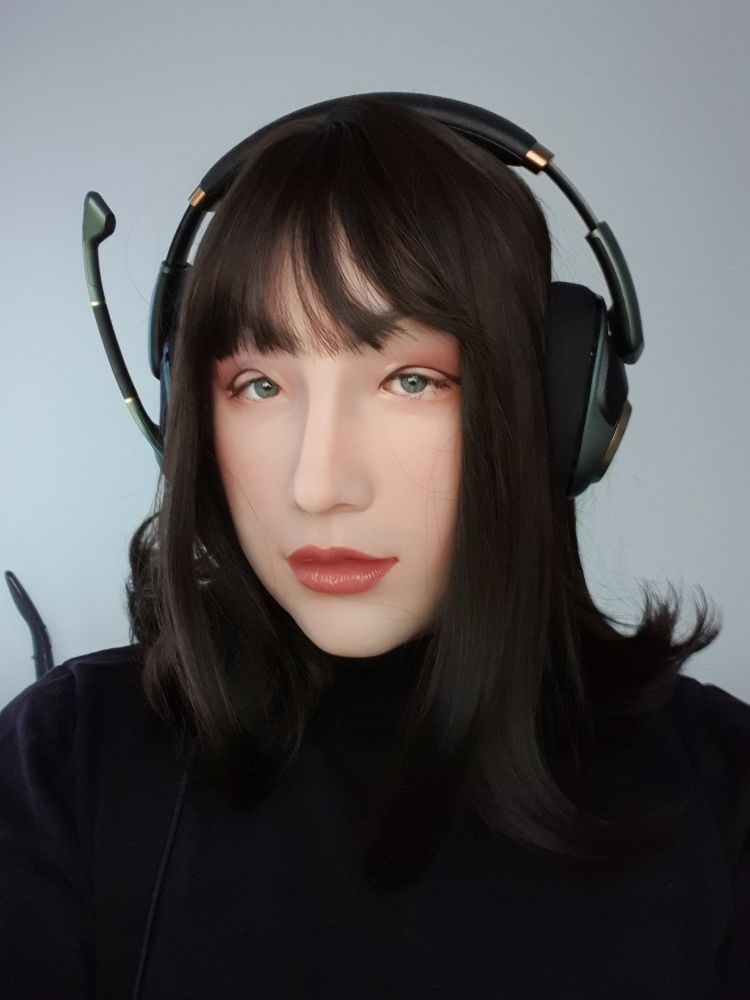 A person wearing a silicone mask of a girl with asian eyes with black hair. The person wears a green headset and a black sweatshirt.