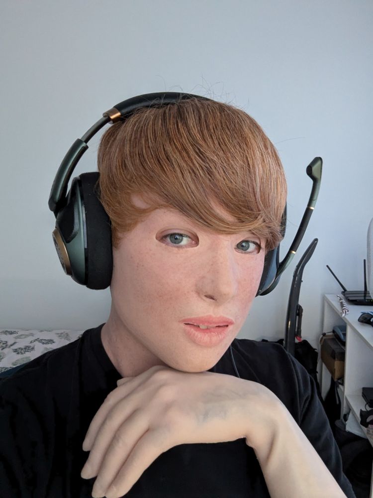 A portrait of a person in a realistic silicone mask of a light-skinned Girl with short blonde hair and light-skinned realistic silicone gloves, wearing a black T-shirt and a Gaming-Headset in a bright room.