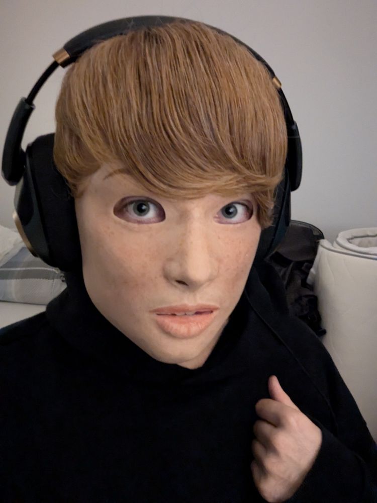 Portrait of a person wearing a realistic silicone mask of a light-skinned freckled girl with a short blonde wig wearing headphones and a black hoodie.