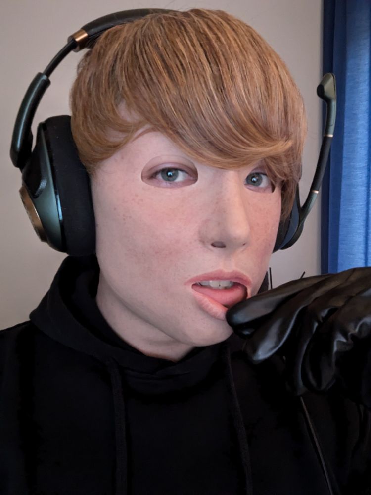 A portrait of a person wearing a realistic silicone mask of a freckled Girl with short blonde Hair, wearing a black hoodie tugging at the lip of the mask with a hand in a black latex glove.