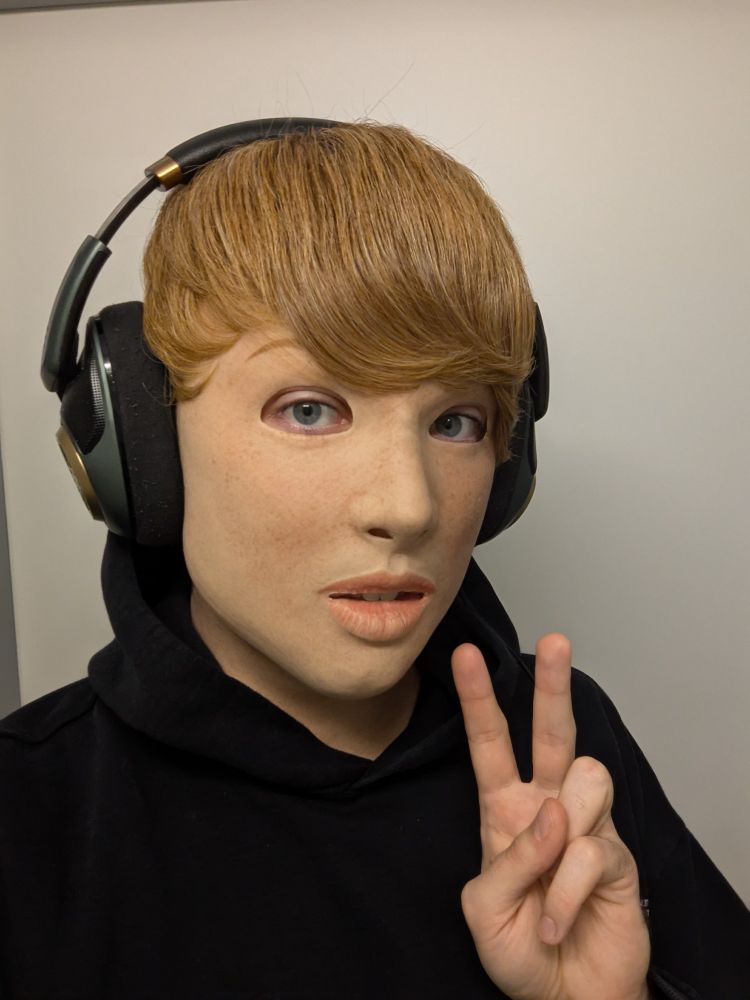 Portrait of a person wearing a realistic silicone mask of a light-skinned freckled girl with a short blonde wig wearing headphones and a black hoodie.