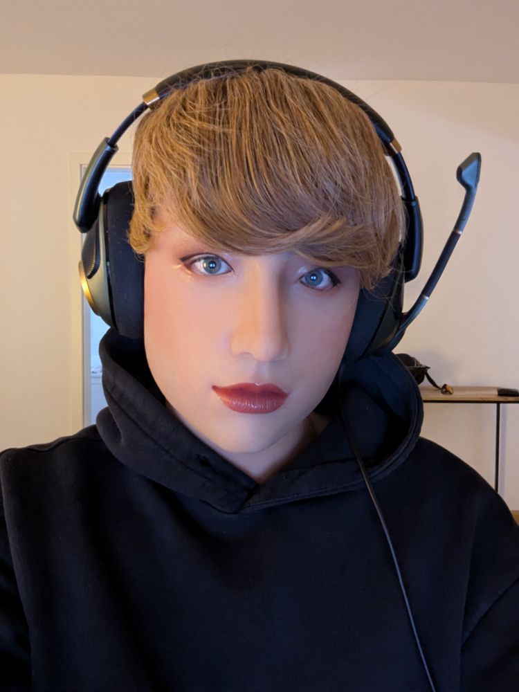 A person wearing a relatively realistic silicone mask of an eurasian looking girl with pale skin, slight makeup and a blonde Wig. The person wears a Headset and a black Hoodie.