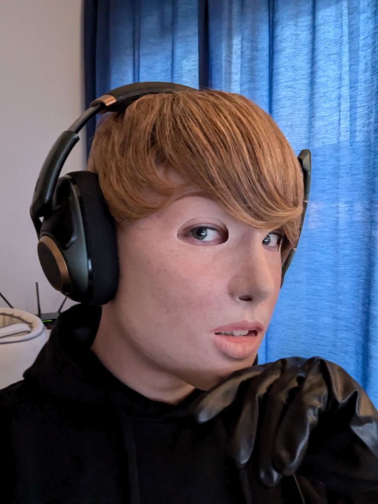 A portrait of a person wearing a realistic silicone mask of a freckled Girl with short blonde Hair, wearing a black hoodie and holding a hand in a black latex glove to the face.