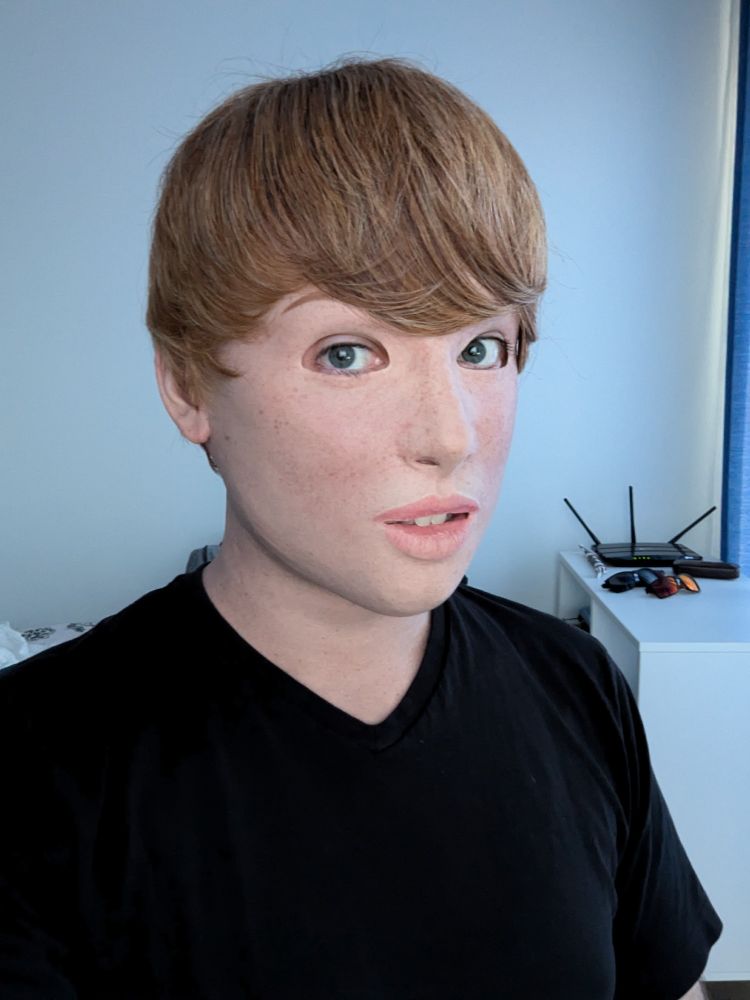 A portrait of a person in a realistic silicone mask of a light-skinned Girl with short blonde hair, wearing a black T-shirt in a bright room.