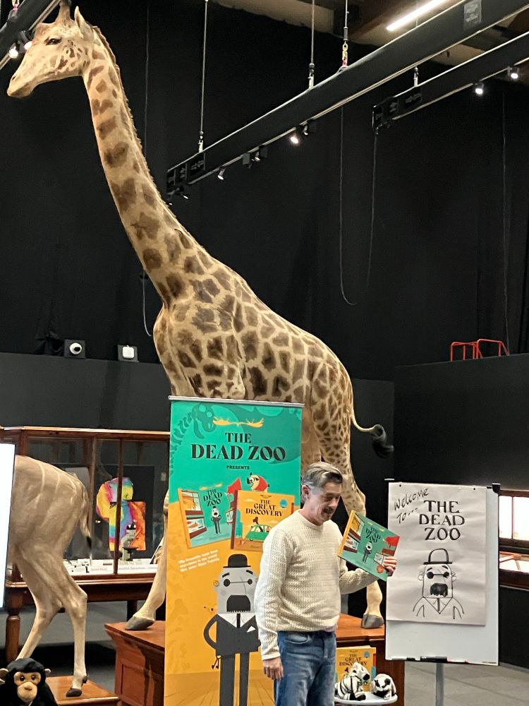 Author and illustrator Peter Donnelly beside Spoticus the giraffe with banner promoting The Dead Zoo book 