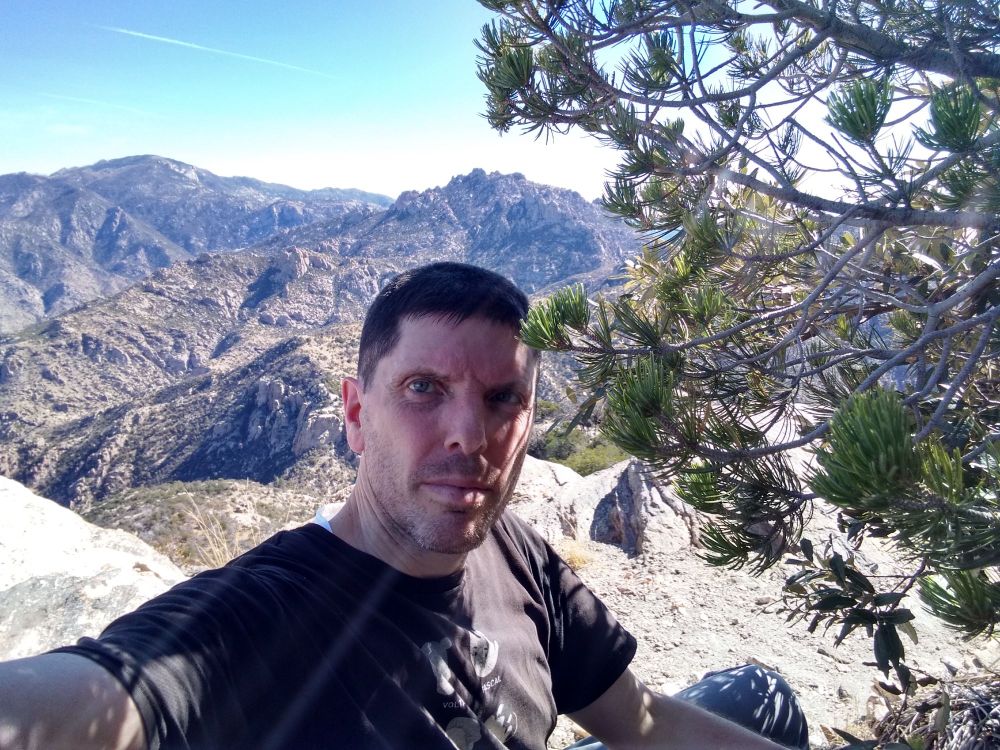 Tom next to tree near cliff edge with rugged mountains behind.