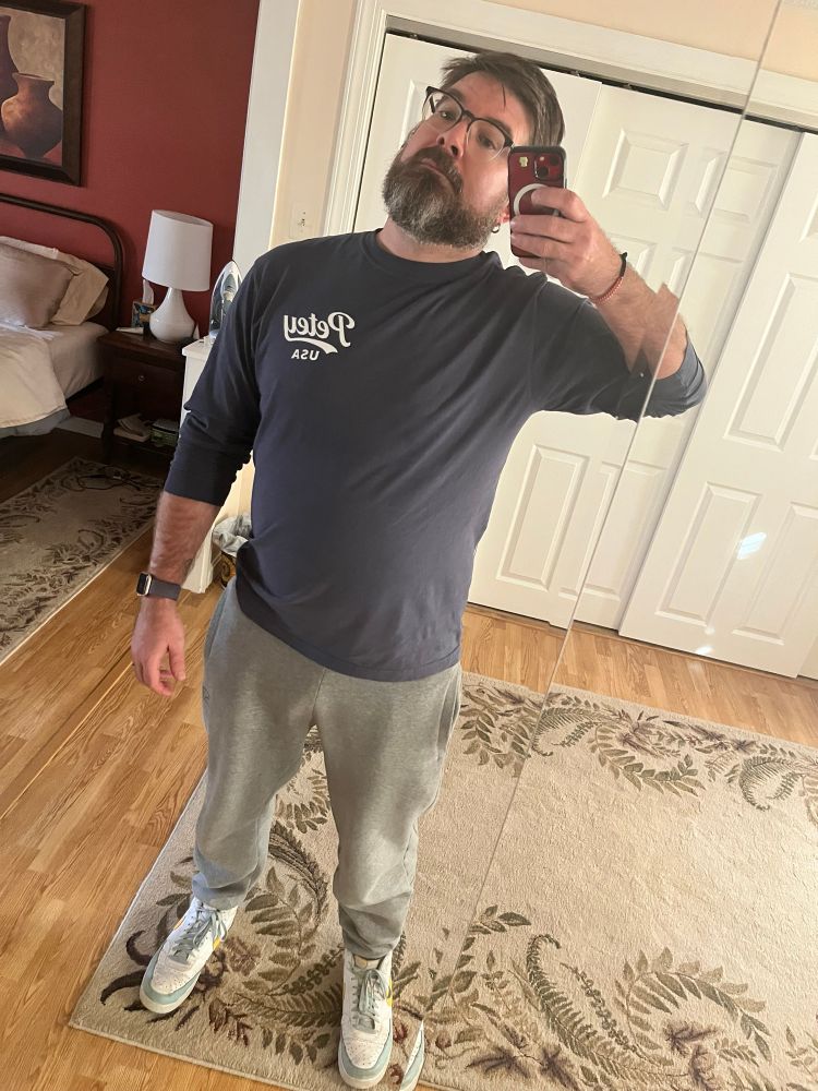 A ma wearing sweatpants gray and a blue shirt taking a mirror selfie. 