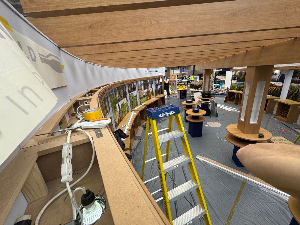 A view across an exhibition stand from up a ladder looking at the progress of the build near complete. On the left some vinyl lettering in the process of being stuck to the wall above a wooden header with light fittings in. Off in the distance are various display stand tables with photographic images of vineyards and winemakers. A step ladder is in the scene also!