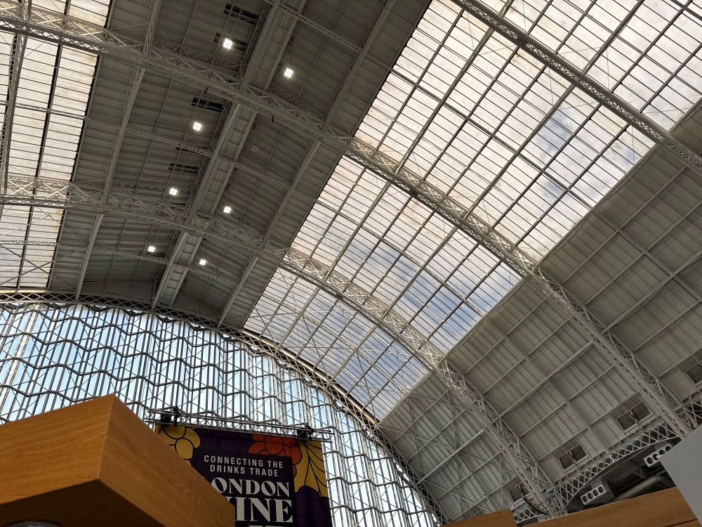 A view up inside from an exhibition stand floor to the arched partly glazed roof of the Kensington Olympia Grand Hall where the clouds and blue sky can be seen through the many many panes of glass above us. A hint of the hanging sign saying London Wine Fair below 