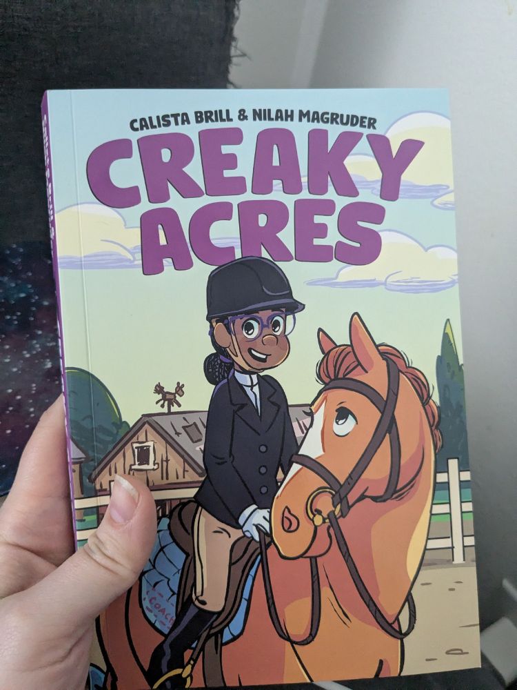 Photograph of graphic novel Creaky Acres by Calista Brill and Nilah Magruder. The cover art is of main character Nora, a happy Black girl in her competition-ready dark jacket and riding helmet sitting on her beloved horse Hay Fever.
