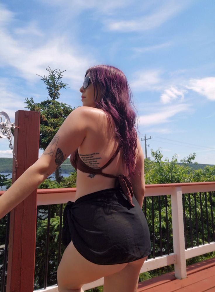 Skye standing on a patio on a clear summer day, she's looking out at the ocean, head and body turned back on to the camera, wearing a brown bikini and a sheer black cover-up on the bottom