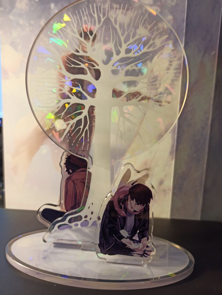 A photo of an acrylyc standee for S classes that i raised made out of three piecea. In the middle is the white tree. Behind it is Han Yoohyun with his back tobthe viewer, hunched forwards. At the front is Han Yoojin with a single memory pearl in his hands.