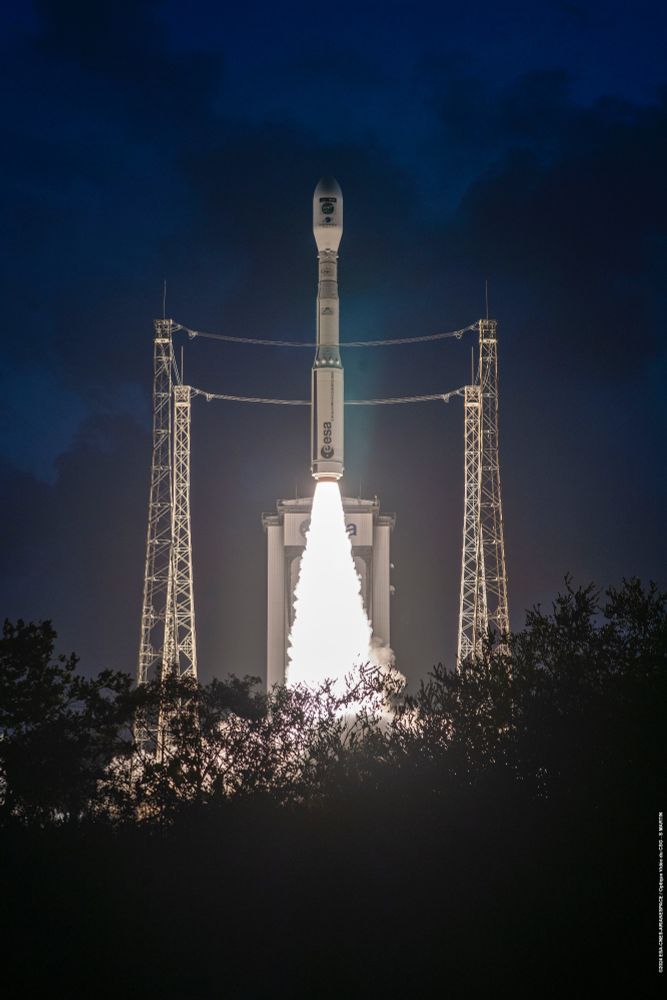 Vega rocket clears launch pad , with four lightning protection towers at sides, dark night skies behind.