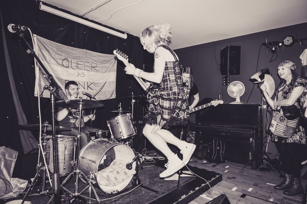 White blonde woman with electric guitar jumping in front of stage with man at drum kit on