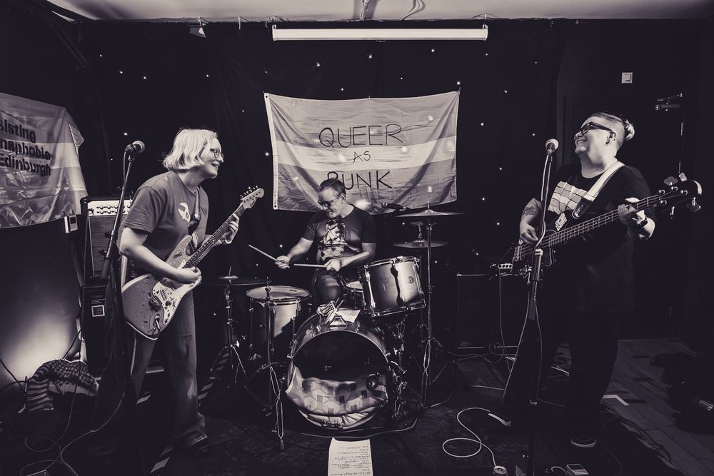 Band on Stage, Trans flag at back. L to R: white woman playing guitar, white man playing drums, white person playing bass. 