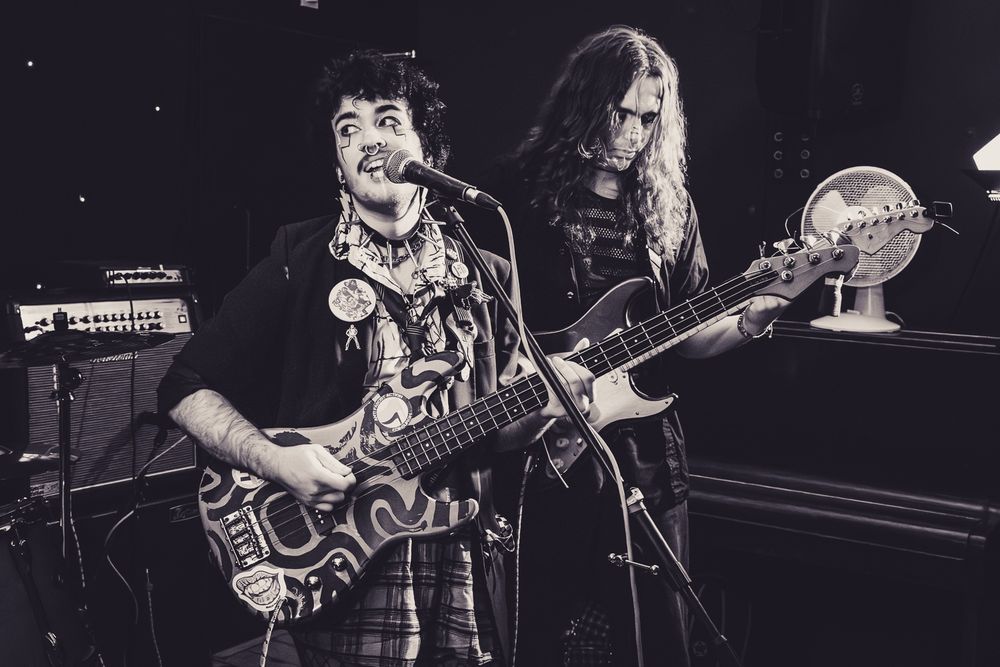 Masc person, pale skin, short hair, wearing makeup, playing bass. Person with long hair behind, playing guitar. 