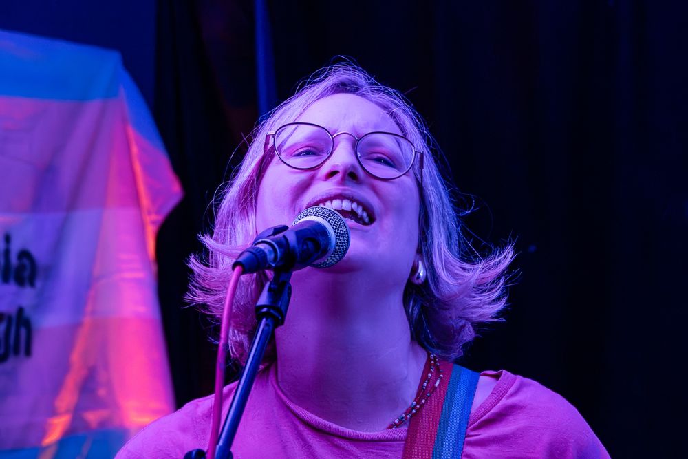 Head and shoulders white woman with glasses and chin length hair sings into microphone 