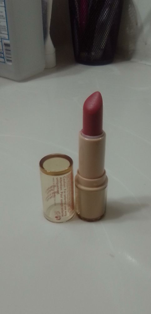 A tube of red lipstick with the product turned up sits on a white faux marble bathroom counter next to its lid. A bottle of isopropyl alcohol & a cup holding toothbrushes can be seen behind the lipstick.