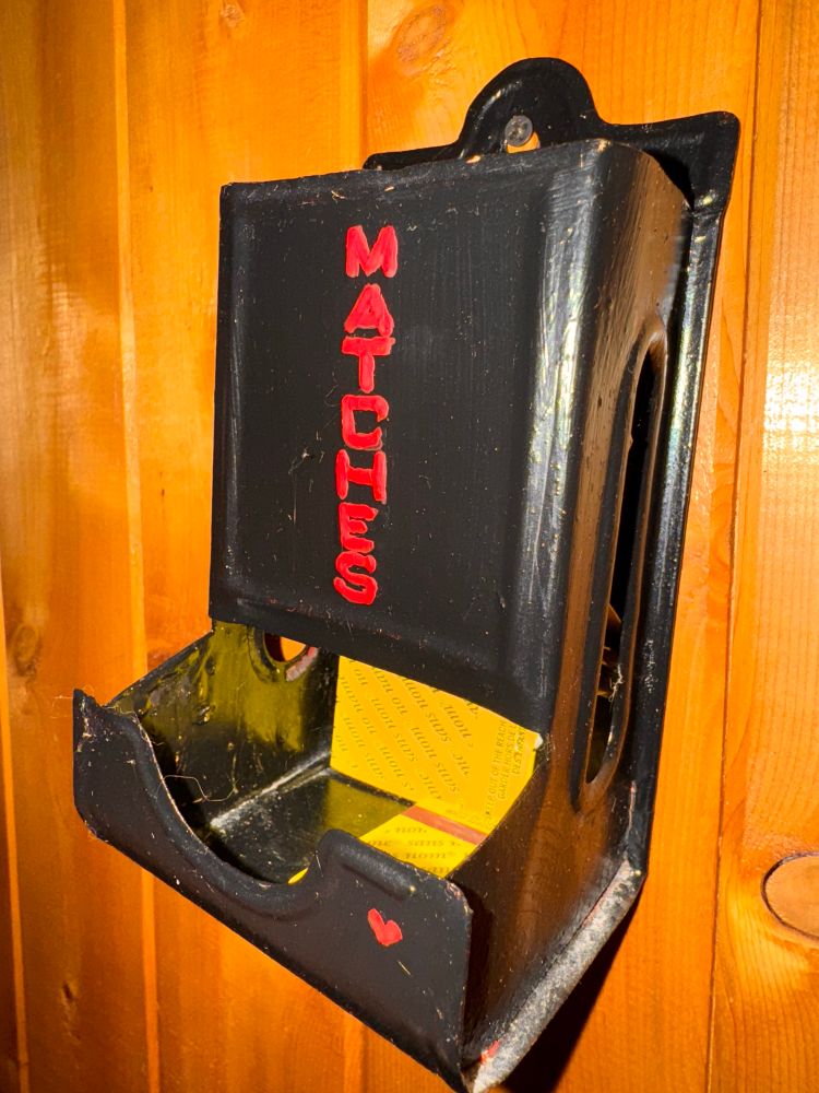 A metal box, painted shiny black is nailed to a wall. The word matches is printed on the front. A few matchboxes are visible in the open drawer at the bottom. 