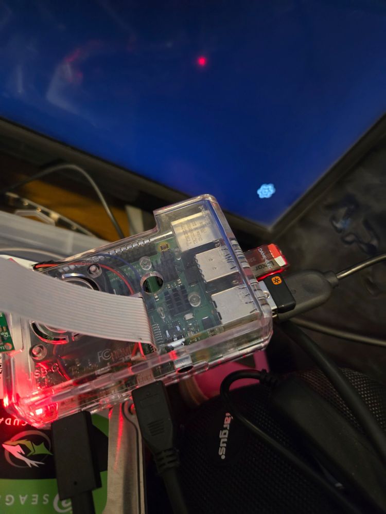 A Raspberry Pi 4 in a clear case with various connections including a USB mouse receiver, keyboard, and USB micro SD card reader with a card inside. The Pi also has a camera connected with a camera ribbon attached to the board.