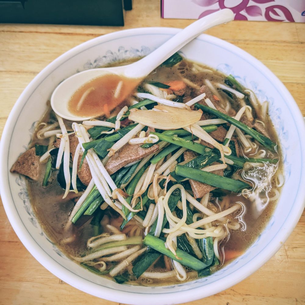 中華風のラーメンの上に、もやし、ニラ、玉ねぎ、レバーなどの具材がたっぷりのっている醤油ベースのスープ麺の写真。白いレンゲ付き。