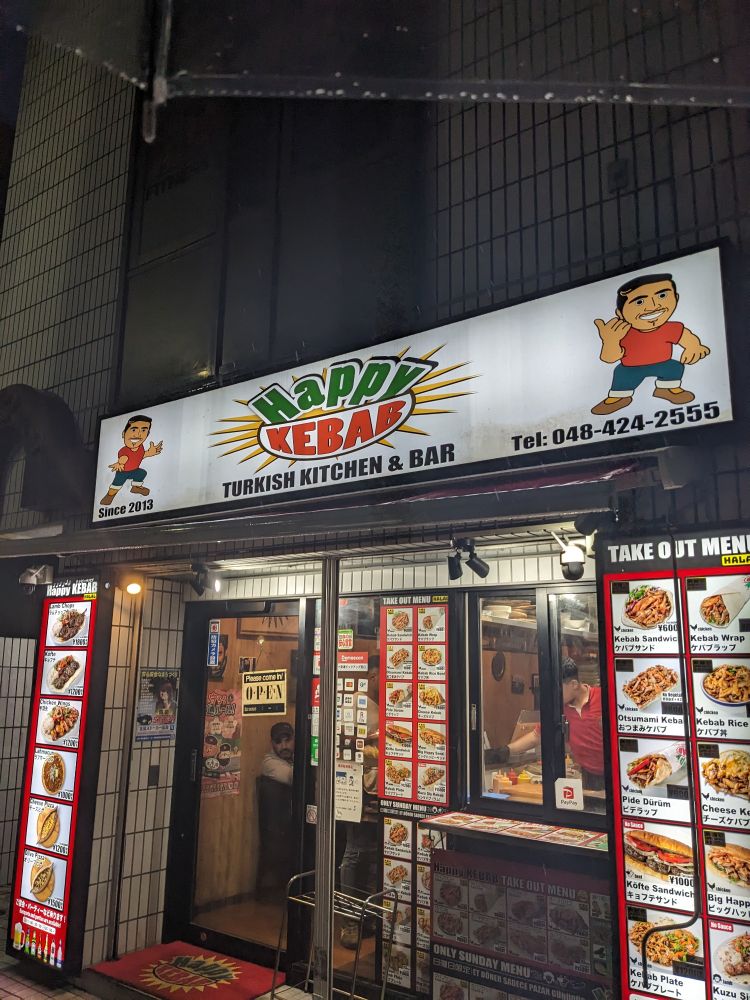 ハッピーケバブというトルコ料理店の外観。看板にキャラクターと「HAPPY KEBAB」「TURKISH KITCHEN & BAR」「Since 2013」と記載。メニュー写真多数。