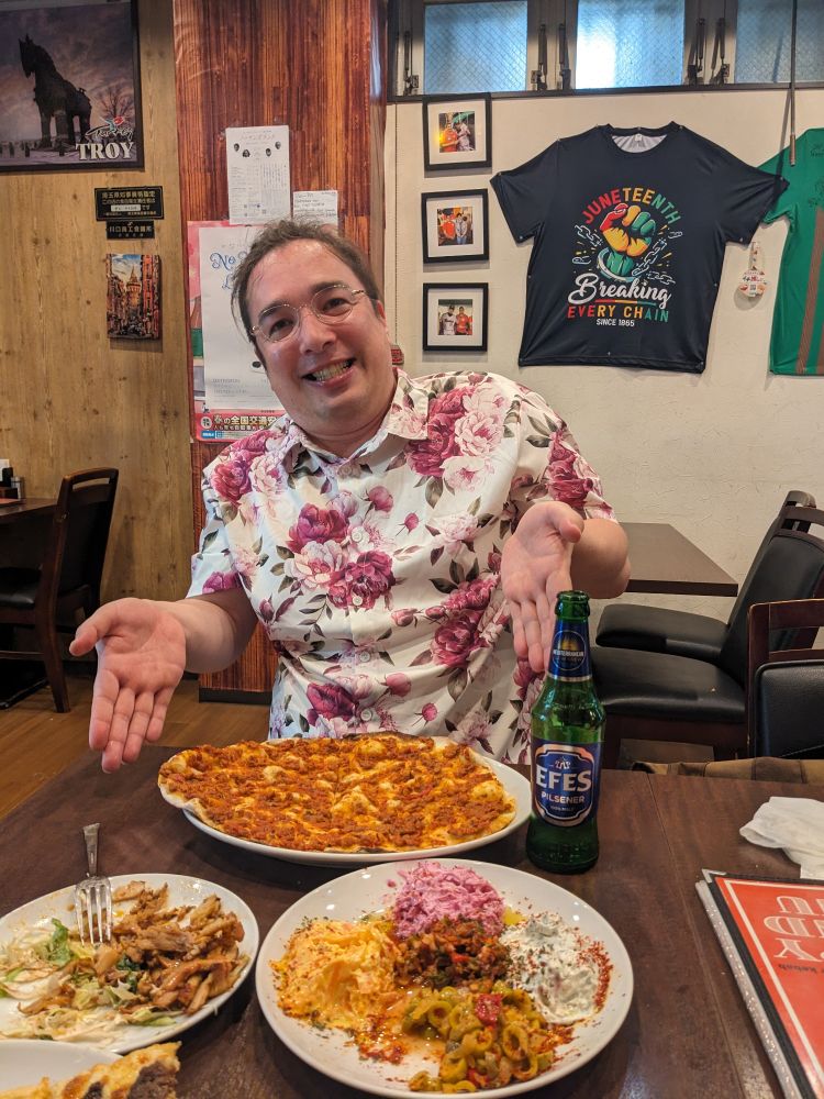 花柄のシャツを着た男性がレストランで笑顔を見せながら料理を紹介しており、テーブルにはピザ、トルコ料理の盛り合わせ、EFESビールが置かれている。壁には「JUNETEENTH Breaking Every Chain SINCE 1865」と書かれたTシャツが飾られている。
