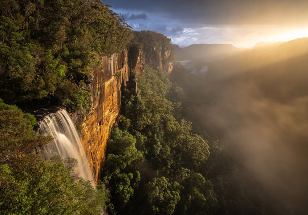 A waterfall is on the bottom left, lit by the sun, Mt Carrialoo is silhouetted in the distance, fog and rain fill the valley between.