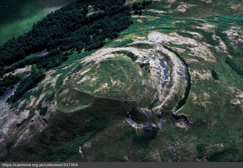 A rather dark aerial photo of the fort covered in a thin dusting of snow. The snow highlights the ramparts and ditches and can be seen as particularly massive toward the right of the image. In the enter there is a central enclosure and to the left a smaller annex can be seen. On the steeper slopes to the top there is a plantation of pine trees.