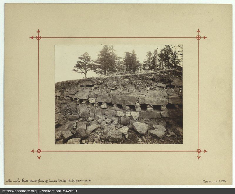 The image shows an old black and whit photo, mounted on sepia coloured paper, with a thin red line, with arrows at each corner, framing the photograph. The photo shows the excavated rampart with a round ended shovel lent against it. Along the line of the dry stone wall there are holes where wooden beams where once located. There is rubble in the foreground, a layer of turf on top of the rampart and trees in the background.