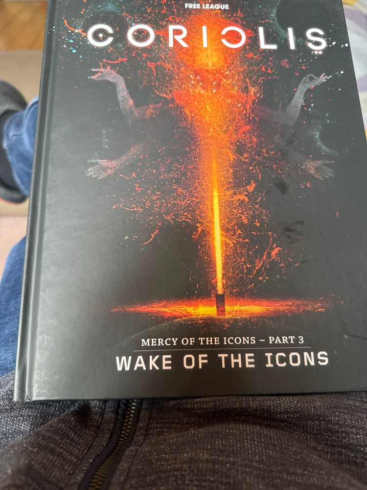 Foto vom englischen Buch “Coriolis - Mercy of the icons - part 3 - wake of the icons”, auf dem Cover sieht man rote Flamen als Strahl und Partikel die eine vierarmige Gestalt mit Kopf in geometrischen Formen andeutet aus der Dunkelheit des Hintergrunds