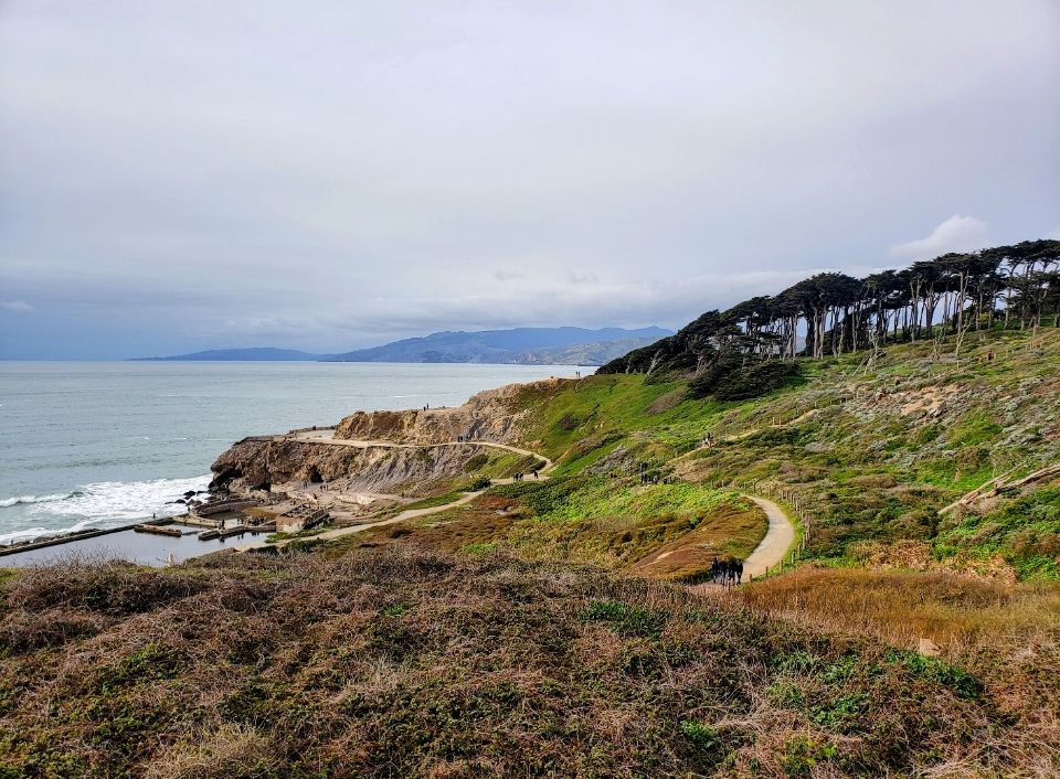 Landscape view of winding trail along Pacific Ocean with cypress trees in middle distance and mountains in the background.
