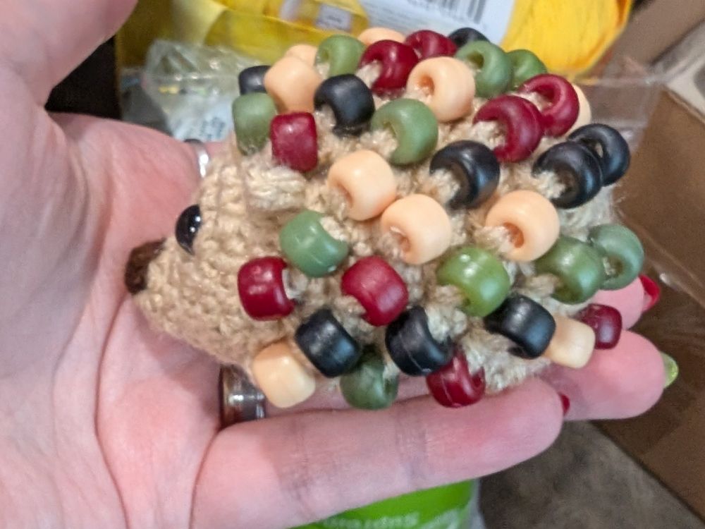 Side view of a crocheted hedgehog with beads for spines. The hedgehog body is a cream color, while the beads are red, black, green, and soft yellow.