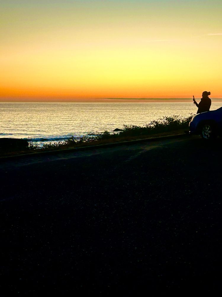 Silhouette  of a person holding up a cell phone as they look out over the ocean and the peach colored sky at sunset.