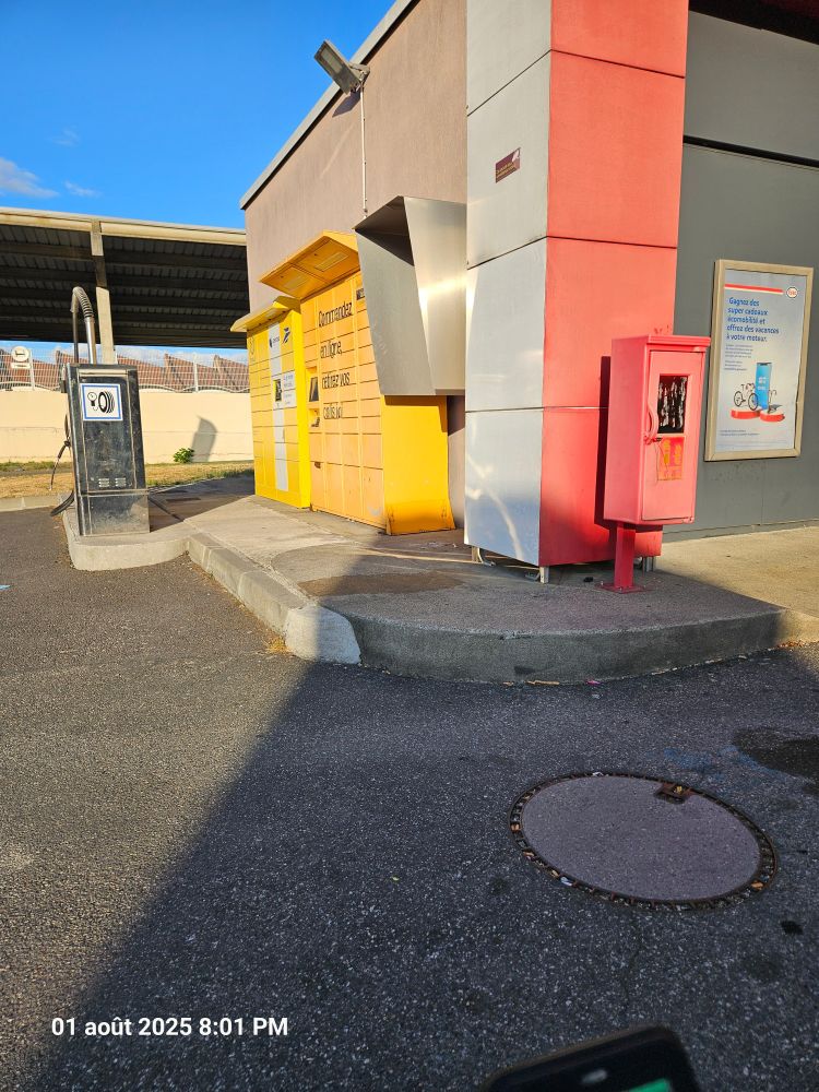 Station-service Esso Express avec casiers de consignes Pickup jaunes et aspirateur posés sur un trottoir surélevé. Aucun abaissement du trottoir n'est visible, empêchant l'accès aux personnes en fauteuil roulant. Les casiers Pickup sont placés à différentes hauteurs sans casiers dédiés aux personnes à mobilité réduite situés entre 90 cm et 1,30 m comme l'exige la réglementation. Le sol présente une plaque d'égout circulaire sur l'asphalte.