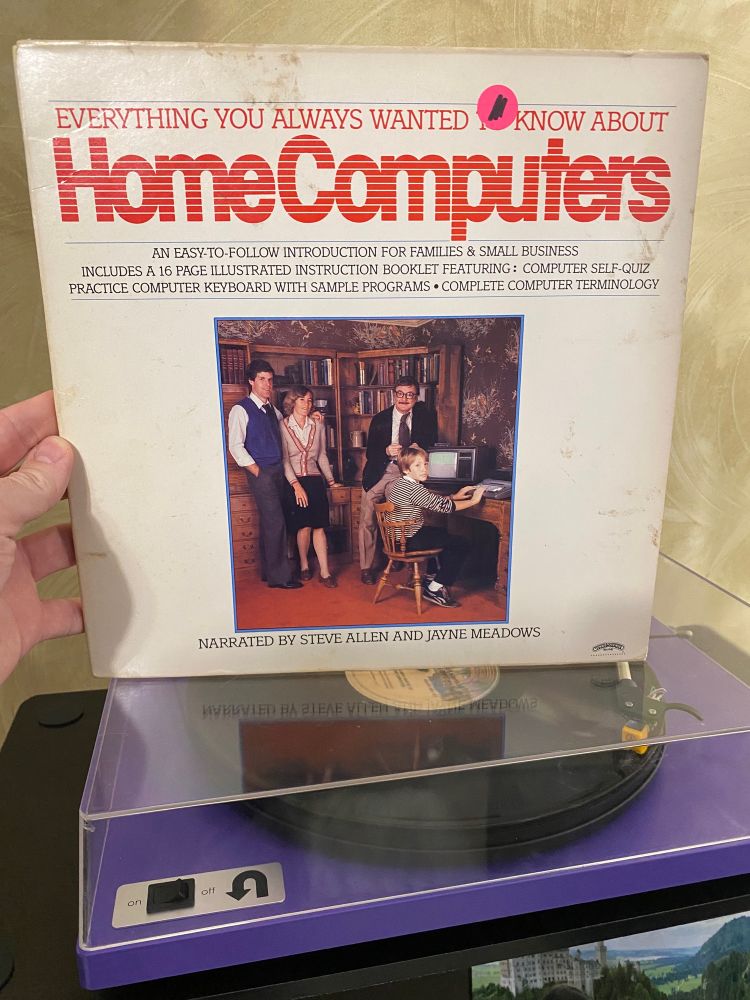 a record sleeve which reads “Everything You Always Wanted to Know About Home Computers: An easy to follow introduction for families & small business / Includes a 16 page illustrated instruction booklet featuring: computer self-quiz, practice computer keyboard with sample programs, complete computer terminology. Narrated by Steve Allen and Jayne Meadows.” and contains an extremely 1980s photo of a couple standing at left of frame in front of a bookshelf, a man in coat and tie with thick frame glasses and a mustache with one leg up on a desk, and a child with an uncomfortable expression seated at the desk with his hands on the keyboard of a now-ancient computer. The sleeve is being held atop a purple turntable upon which the record is playing.
